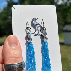 May include: Dangle earrings featuring silver-tone hooks, two small circular iridescent shell accents, and long, bright blue beaded fringe tassels. The earrings are displayed on a white card with a black raven logo and the text Crow Clan Beadwork.