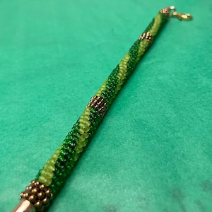 May include: A tubular beaded bracelet featuring a spiral pattern of lime green and forest green glass beads. The design includes metallic bronze accent beads at intervals and is finished with a gold-toned lobster claw clasp at one end.