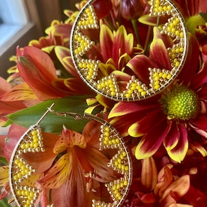 May include: Two gold hoop earrings with yellow beaded geometric designs. The earrings are displayed against a backdrop of red, orange, and yellow flowers, creating a vibrant and eye-catching composition. The earrings are a stylish accessory.