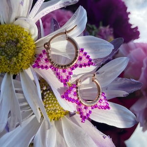 May include: A pair of gold hoop earrings with a geometric design made of small pink and white beads. The earrings are displayed on a white daisy with a yellow center, with a blurred background of purple and pink flowers.