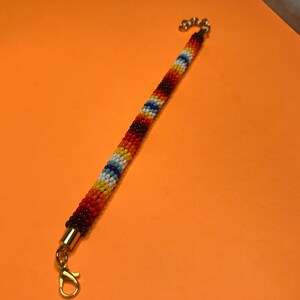 May include: A beaded bracelet with a cylindrical design. The bracelet features a pattern of red, orange, yellow, white, blue, and black beads. Gold-colored metal caps and a clasp complete the bracelet.