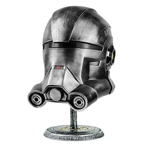 May include: A silver-colored helmet with a weathered finish, featuring a visor and a detailed design. The helmet is displayed on a black stand with a decorative base.