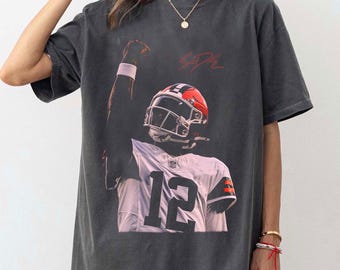 May include: Dark gray t-shirt featuring a football player in a white and brown uniform with the number 12. The player is wearing a helmet and raising their arm. The word "SADIE" is written in red script.