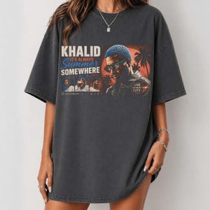 May include: A charcoal gray oversized t-shirt featuring a graphic print of Khalid. The design includes the text 'KHALID IT'S ALWAYS SUMMER SOMEWHERE TOUR', 'LIVE IN YOUR CITY 2024', and three small portrait photos of the artist against a sunset background.