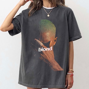 May include: Dark gray t-shirt featuring a graphic of a person with green hair and the word "blond" in white. The image is a close-up of the person's face and hand. The shirt has short sleeves and a crew neck.