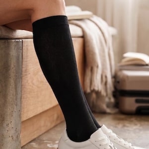 May include: A person wearing black knee-high socks and white sneakers with brown soles. The sneakers are laced up and appear to be made of leather. The background includes a wooden bench, a blanket, and a suitcase.