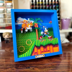 Op de afbeelding: Een blauw ingelijst diorama met een gepixelde scène uit het videospel Sonic the Hedgehog. Het diorama bevat Sonic die rent, Tails die vliegt en een palmboom. Het diorama is gemaakt van plastic kralen en is ongeveer 25 cm breed.