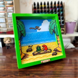 Peut inclure: Un cadre photo vert avec une scène de plage pixelisée à l'intérieur. La scène représente deux personnages assis sur une bûche sous des palmiers. Le cadre a une petite plaque en bois avec le texte "Love & Marine" dessus.