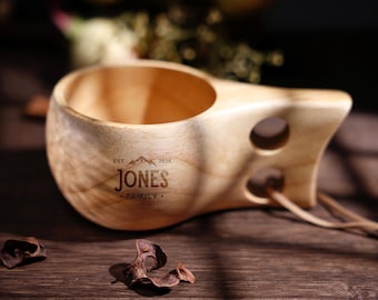 Może przedstawiać: Drewniany kubek kuksa z uchwytem i dwoma otworami. Kubek jest grawerowany napisem "JONES FAMILY" i rokiem "2025" wraz z grafiką górską. Kubek wykonany jest z jasnego drewna i stoi na ciemnej drewnianej powierzchni.