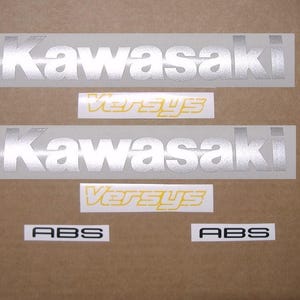 Op de afbeelding: Een verzameling Kawasaki motorfietsstickers. De set bevat twee zilveren Kawasaki logo stickers, twee geel-witte Versys stickers en twee ABS stickers met zwarte tekst op een witte achtergrond.