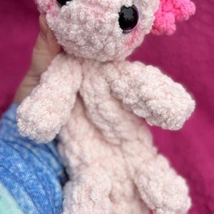 May include: A pink and white crocheted axolotl plush toy with large black eyes. The axolotl has pink feathery gills and a pink tail.