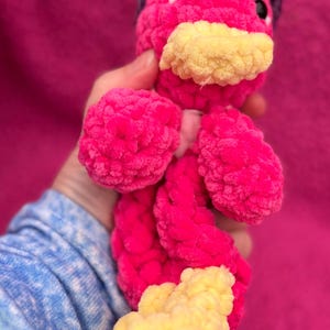 May include: A pink crocheted plush toy with a purple and white crocheted hat. The toy has yellow crocheted feet and a yellow crocheted beak.