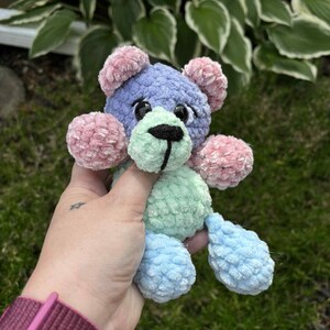 May include: A handmade crochet teddy bear in pastel colors. The bear features a blue head, pink ears and cheeks, a green muzzle, and blue feet. The bear has a black stitched nose and eyes. The soft toy is held in a hand.