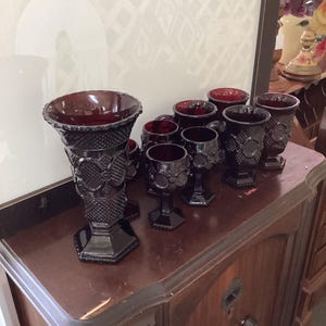 Puede incluir: Una colección de cristalería roja rubí vintage, que incluye un jarrón grande y varias copas y vasos más pequeños. La cristalería presenta un patrón detallado en relieve y se encuentra sobre una superficie de madera oscura. La iluminación resalta el color intenso y los diseños intrincados.