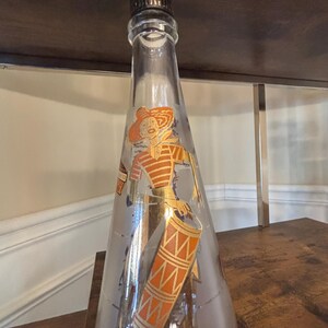 May include: Clear glass bottle with a decorative image of a person in a hat and a drum. The bottle has a dark, textured stopper. The image on the bottle is in gold and orange. The bottle is on a wooden surface.