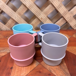 May include: A set of four stackable mugs in pastel colors including light blue, dusty pink, soft grey, and periwinkle blue. Each mug features a smooth matte finish, a cylindrical body with a distinct horizontal ridge, and a small square handle.