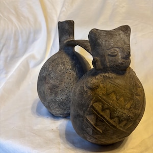 Może przedstawiać: Ciemnoszare naczynie ceramiczne z dwiema połączonymi kulistymi komorami. Jedna komora zakończona jest pionową szyjką, a druga ma kształt stylizowanej głowy kota z wyrytymi geometrycznymi wzorami i małymi kropkami na powierzchni.
