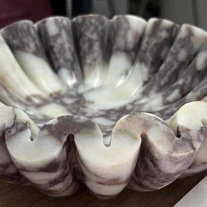 Pu&ograve; includere: Una ciotola decorativa, smerlata, in marmo. La ciotola presenta un motivo marmorizzato bianco e viola. I bordi della ciotola hanno un design ondulato, a forma di conchiglia. La ciotola &egrave; appoggiata su una superficie di legno.