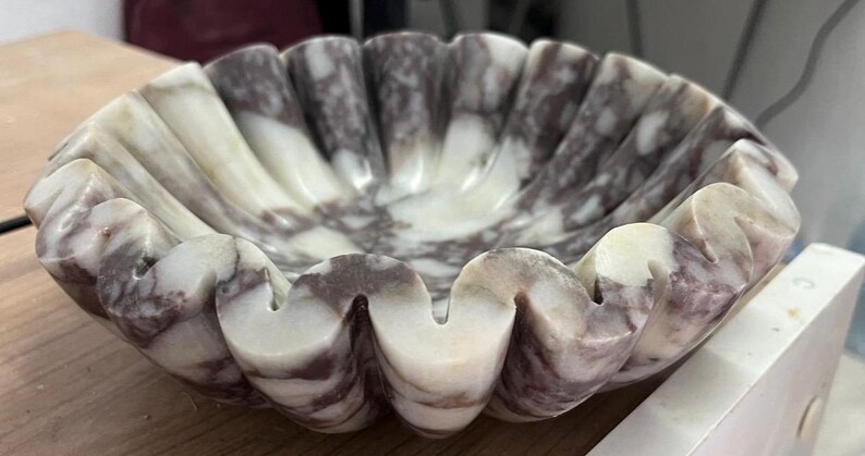 Pu&ograve; includere: Una ciotola decorativa, smerlata, in marmo. La ciotola presenta un motivo marmorizzato bianco e viola. I bordi della ciotola hanno un design ondulato, a forma di conchiglia. La ciotola &egrave; appoggiata su una superficie di legno.