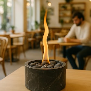 May include: A small, dark grey, cylindrical tabletop fireplace with a marble-like pattern. The fireplace contains dark grey stones and a tall, slender flame. The fireplace is on a light brown wooden table in a cafe setting.
