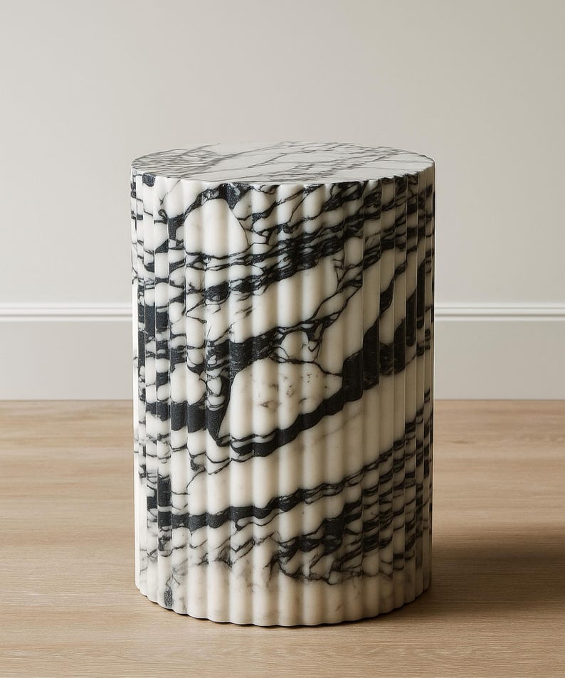 May include: A cylindrical marble side table with a fluted design. The table features a black and white veined pattern. The table is approximately 18 inches tall and 14 inches in diameter, suitable for indoor use.
