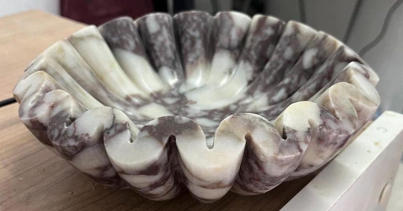Pu&ograve; includere: Una ciotola decorativa in marmo con bordo smerlato. La ciotola presenta un mix di venature bianche e viola scuro. Il design della ciotola ricorda una conchiglia.