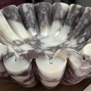 Pu&ograve; includere: Una ciotola decorativa a bordi smerlati realizzata in marmo. La ciotola presenta un bordo ondulato e una tavolozza di colori naturali di venature bianche e grigio-viola. Il design della ciotola ricorda una conchiglia.
