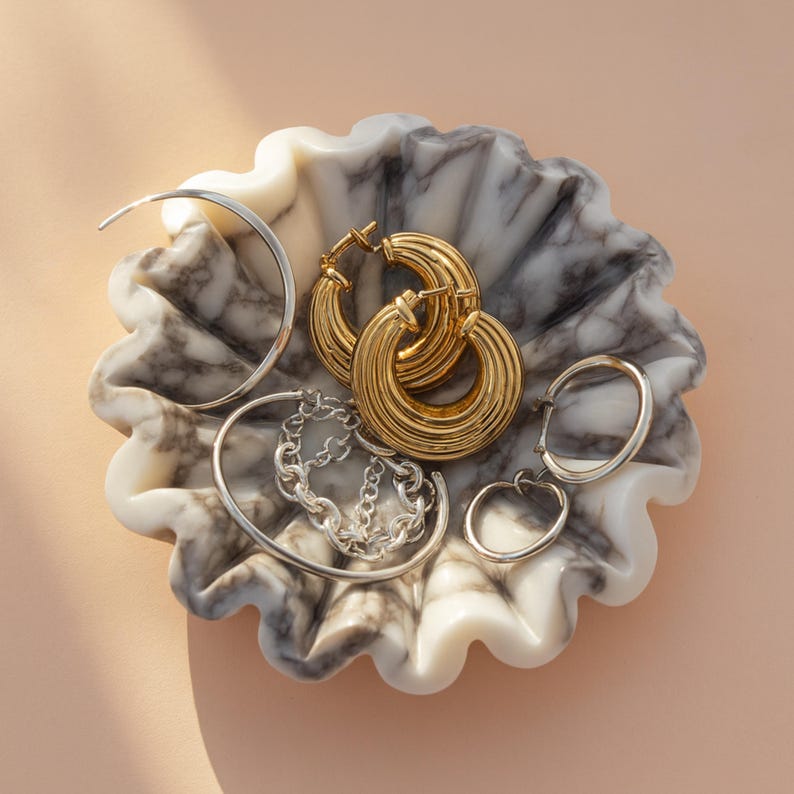 Pu&ograve; includere: Una collezione di orecchini e bracciali in oro e argento esposti in una ciotola decorativa in marmo. Gli orecchini includono grandi orecchini a cerchio dorati, piccoli orecchini a cerchio argentati e un braccialetto a catena d'argento. La ciotola ha un motivo marmorizzato bianco e grigio.