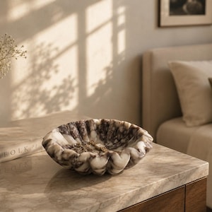 Pu&ograve; includere: Una ciotola decorativa in marmo con bordo smerlato, nei toni del bianco e del marrone. La ciotola &egrave; su una superficie di marmo, con un libro e un vaso di fiori nelle vicinanze. La scena &egrave; illuminata dalla luce naturale.