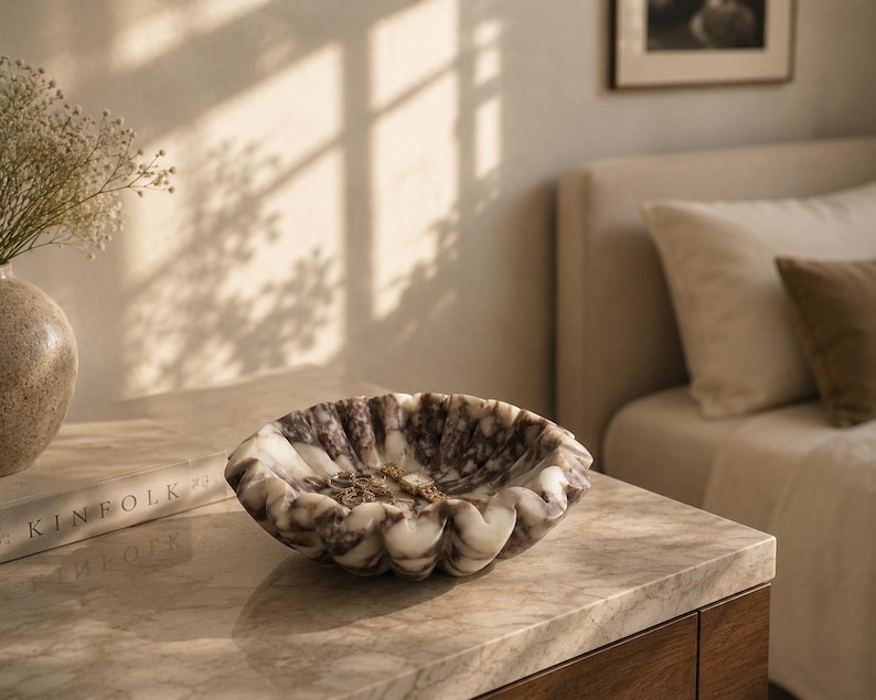 Pu&ograve; includere: Una ciotola decorativa in marmo con bordo smerlato, nei toni del bianco e del marrone. La ciotola &egrave; su una superficie di marmo, con un libro e un vaso di fiori nelle vicinanze. La scena &egrave; illuminata dalla luce naturale.