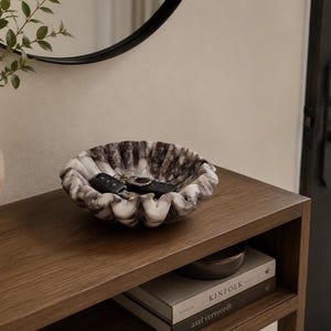 Pu&ograve; includere: Una ciotola decorativa in marmo con bordo smerlato, che mostra una miscela di venature bianche e grigio scuro. La ciotola contiene un set di chiavi ed &egrave; appoggiata su una superficie di legno. Uno specchio rotondo &egrave; visibile sullo sfondo.