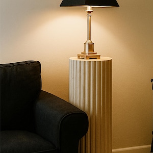 May include: A brass table lamp with a black square lampshade sits atop a fluted, cylindrical wooden pedestal. The lamp is illuminated, casting a warm glow. A black armchair is in the foreground.