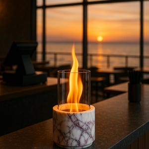 May include: A cylindrical marble tabletop fireplace with a glass enclosure, showcasing a vibrant flame. The base features a white and purple veined pattern. The background shows a sunset over the ocean.