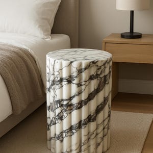 May include: A cylindrical marble side table with a fluted design. The table features a white base with dark gray veining. It is approximately 18 inches tall and is placed on a neutral-colored rug next to a bed.