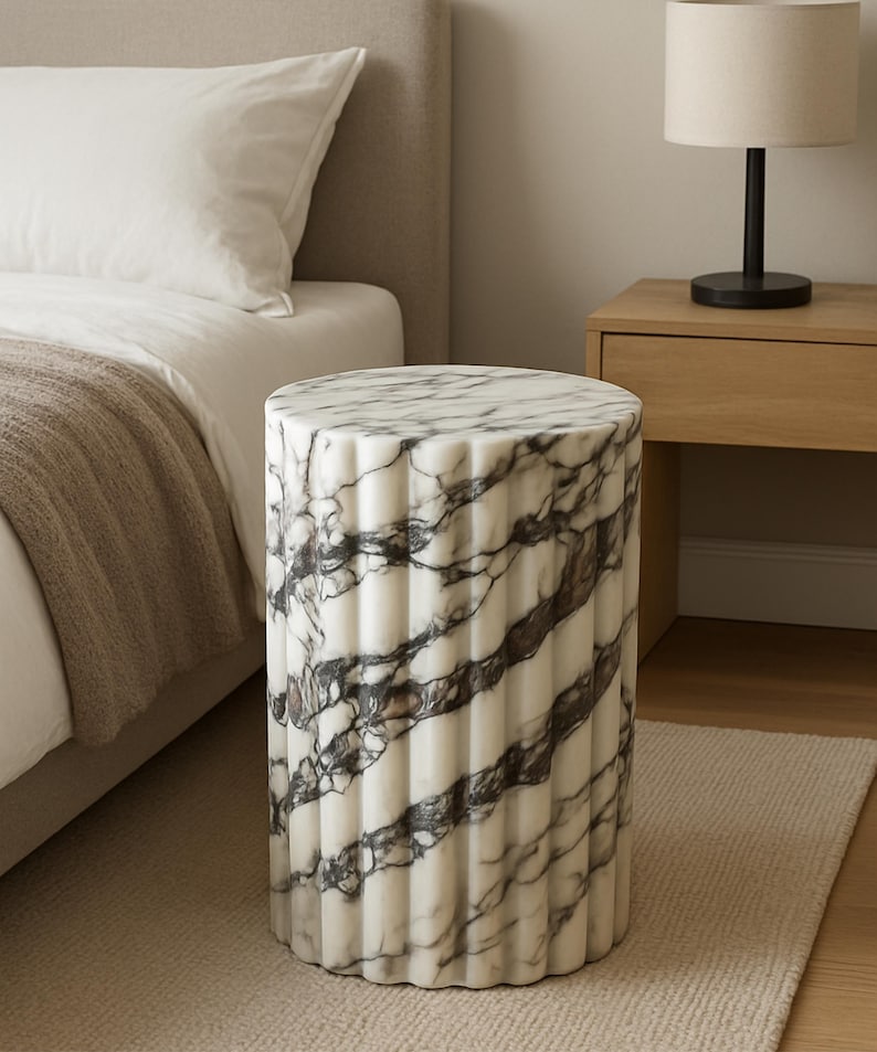 May include: A cylindrical marble side table with a fluted design. The table features a white base with dark gray veining. It is approximately 18 inches tall and is placed on a neutral-colored rug next to a bed.