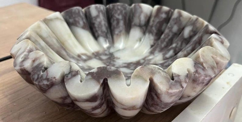Pu&ograve; includere: Una ciotola decorativa smerlata in marmo. La ciotola presenta una combinazione di colori crema e bordeaux con un bordo ondulato e strutturato. L'interno della ciotola presenta un motivo marmorizzato.