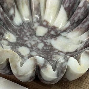 Pu&ograve; includere: Una ciotola decorativa a conchiglia scolpita in marmo. La ciotola presenta un motivo naturale di venature bianche e viola scuro. I bordi sono ondulati, creando un aspetto a conchiglia. La ciotola &egrave; probabilmente usata per l'esposizione o come elemento decorativo.