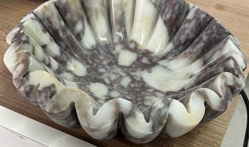 Pu&ograve; includere: Una ciotola decorativa in marmo con bordo smerlato. La ciotola presenta un motivo naturale di venature bianche e grigio-viola. Il design ricorda una conchiglia, con un bordo ondulato e strutturato. Ideale per l'arredamento della casa, circa 25 cm di diametro.