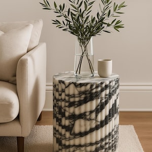 May include: A cylindrical side table with a white and black marble pattern. A clear glass vase with green leafy branches and a small white ceramic cup sit on top. The table is next to a beige sofa.