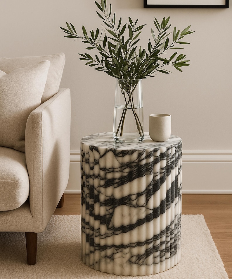 May include: A cylindrical side table with a white and black marble pattern. A clear glass vase with green leafy branches and a small white ceramic cup sit on top. The table is next to a beige sofa.