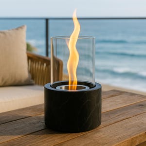 Op de afbeelding: Een cilindrische zwarte marmeren tafelhaard met een glazen omhulsel, met een heldere oranje vlam. De haard staat op een houten oppervlak met een vage oceaan op de achtergrond, wat een warme sfeer creëert. Ongeveer 15 cm hoog.