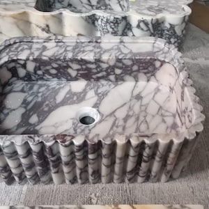 Könnte beinhalten: Ein rechteckiges Waschbecken aus Marmor mit gewelltem Rand. Das Waschbecken hat ein weißes und violett geädertes Muster. Das Abflussloch ist in der Mitte sichtbar. Weitere Marmorwaschbecken sind im Hintergrund.