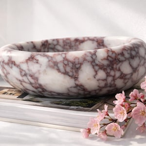 May include: A marble bowl with a white and burgundy veined pattern. The bowl is round and sits on a stack of books, with pink flowers in the foreground. The bowl is a decorative item.