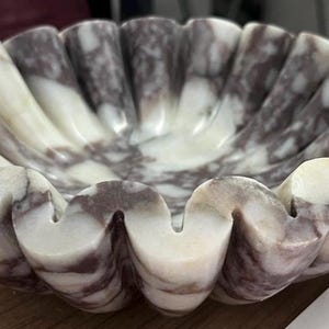 Pu&ograve; includere: Una ciotola decorativa in marmo con bordo smerlato. La ciotola presenta un mix di venature bianche e viola scuro in tutta la pietra. Il design della ciotola ricorda una conchiglia.