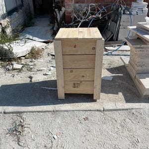 May include: A wooden crate with a light brown finish. The crate has a rectangular shape with four visible horizontal planks and vertical supports at each corner. The crate is outdoors on a concrete surface.