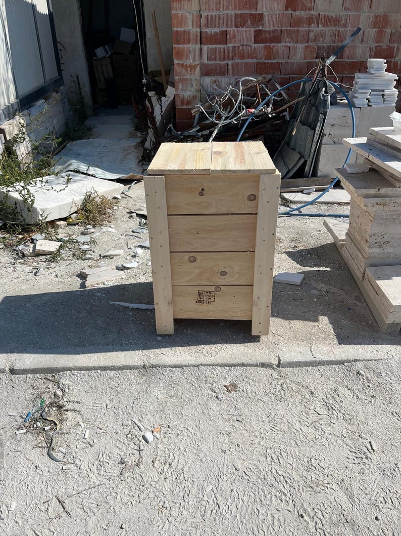 May include: A wooden crate with a light brown finish. The crate has a rectangular shape with four visible horizontal planks and vertical supports at each corner. The crate is outdoors on a concrete surface.