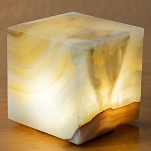 May include: A square onyx lamp, lit from within. The lamp has a warm, yellow and brown colour scheme with a natural, layered pattern. The internal light source creates a soft glow. A decorative home accent.