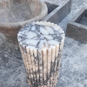 May include: A cylindrical marble pedestal sink with a fluted design. The sink features a white and gray marbled top with a central drain hole. The sides are vertically grooved, creating a textured appearance. Other stone fixtures are in the background.
