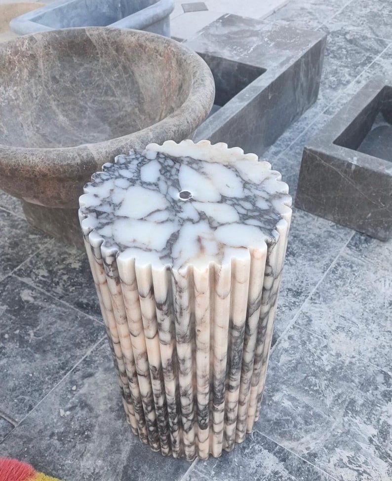 May include: A cylindrical marble pedestal sink with a fluted design. The sink features a white and gray marbled top with a central drain hole. The sides are vertically grooved, creating a textured appearance. Other stone fixtures are in the background.