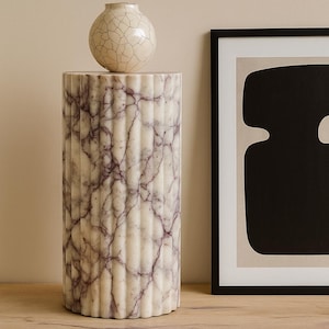 May include: A cylindrical marble pedestal with vertical fluting and a cream and purple veined pattern. A small, round, cream-colored vase with a cracked glaze sits atop the pedestal. A framed abstract art piece is to the right.